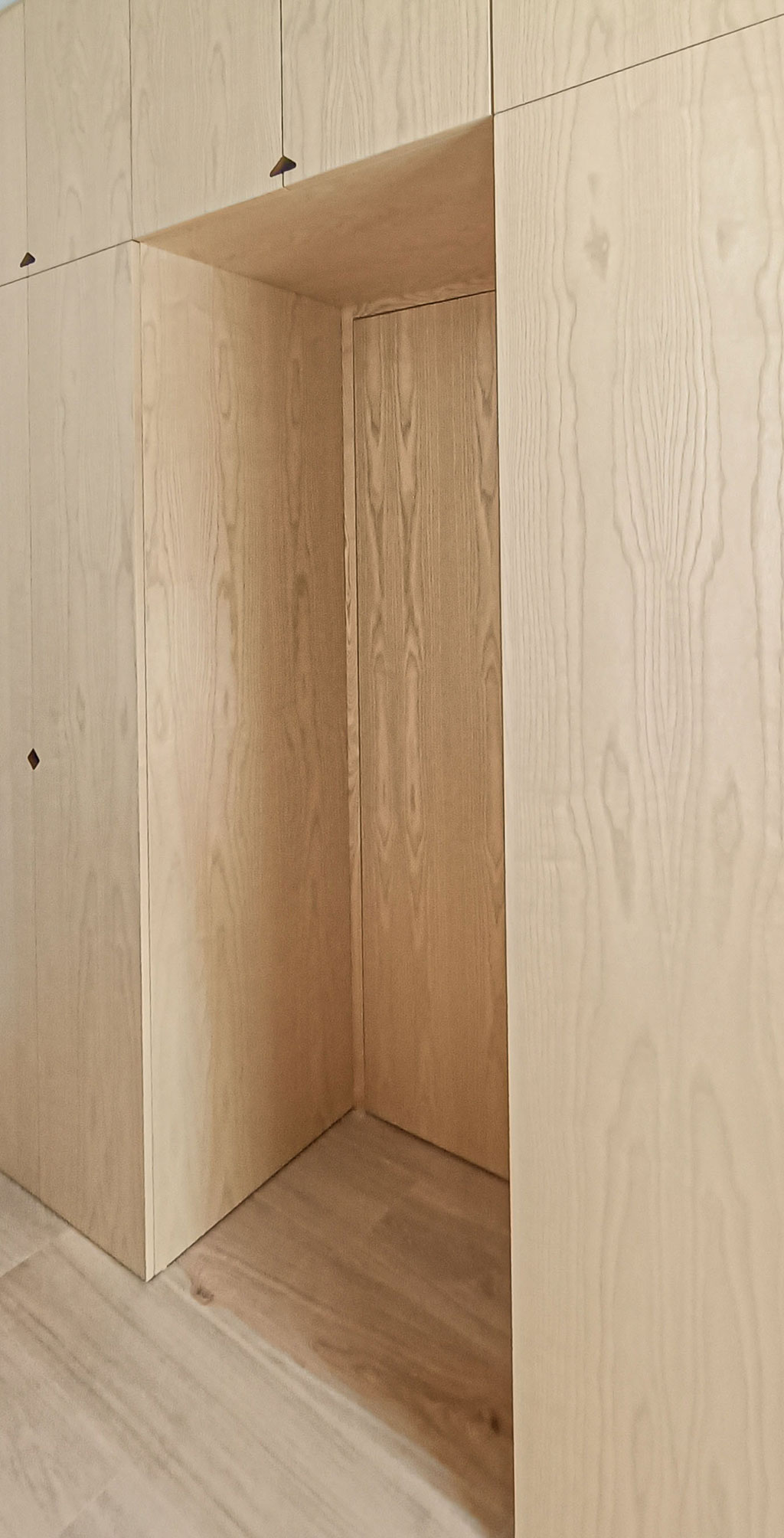 Se plantea como un muro de madera que separa el dormitorio del pasillo, para lo que se integra la puerta  en el propio armario utilizando el mismo tablero, eliminando tapajuntas, bisagras (ocultas) y manilla (oculta de cierre magnético)